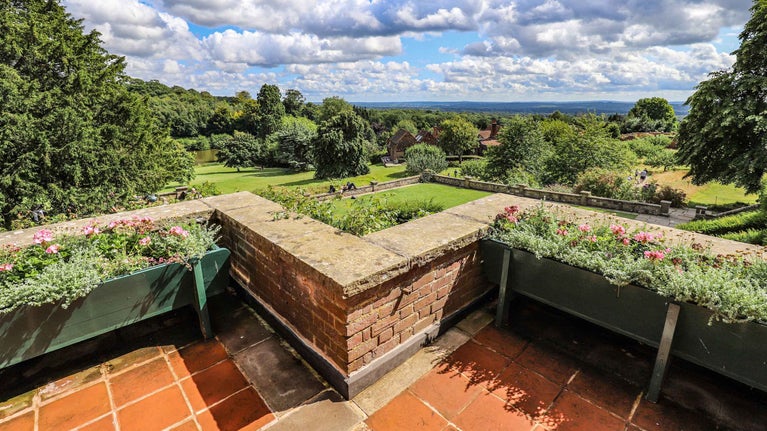 View from the terrace at Chartwell, Kent. The terrace wall is lined with planters of flowers and shrubs, below the terrace the garden can be seen stretching into the distance and the countryside beyond. The summer blue skies are filled with light fluffy clouds.